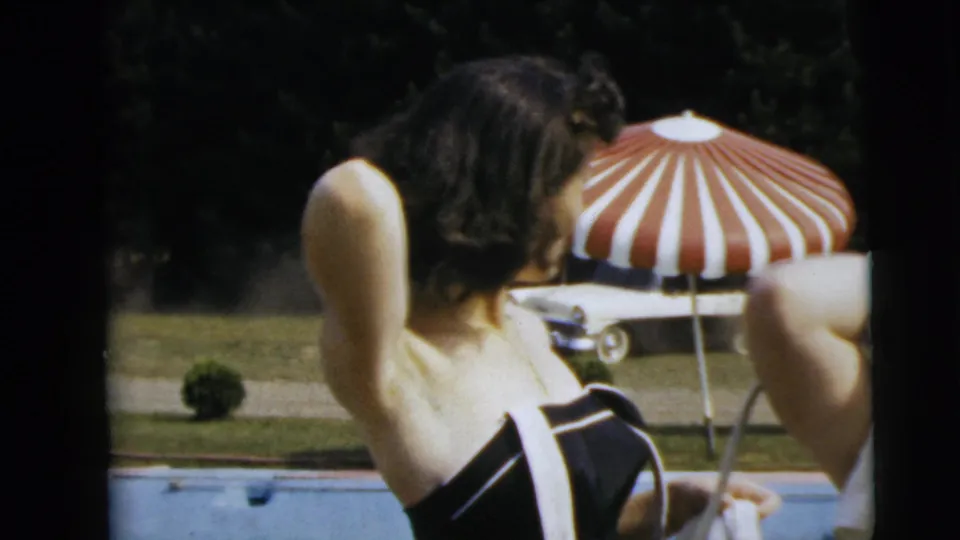 Two adults are partly framed at a backyard pool, with a red-and-white striped umbrella and a period car in the background.