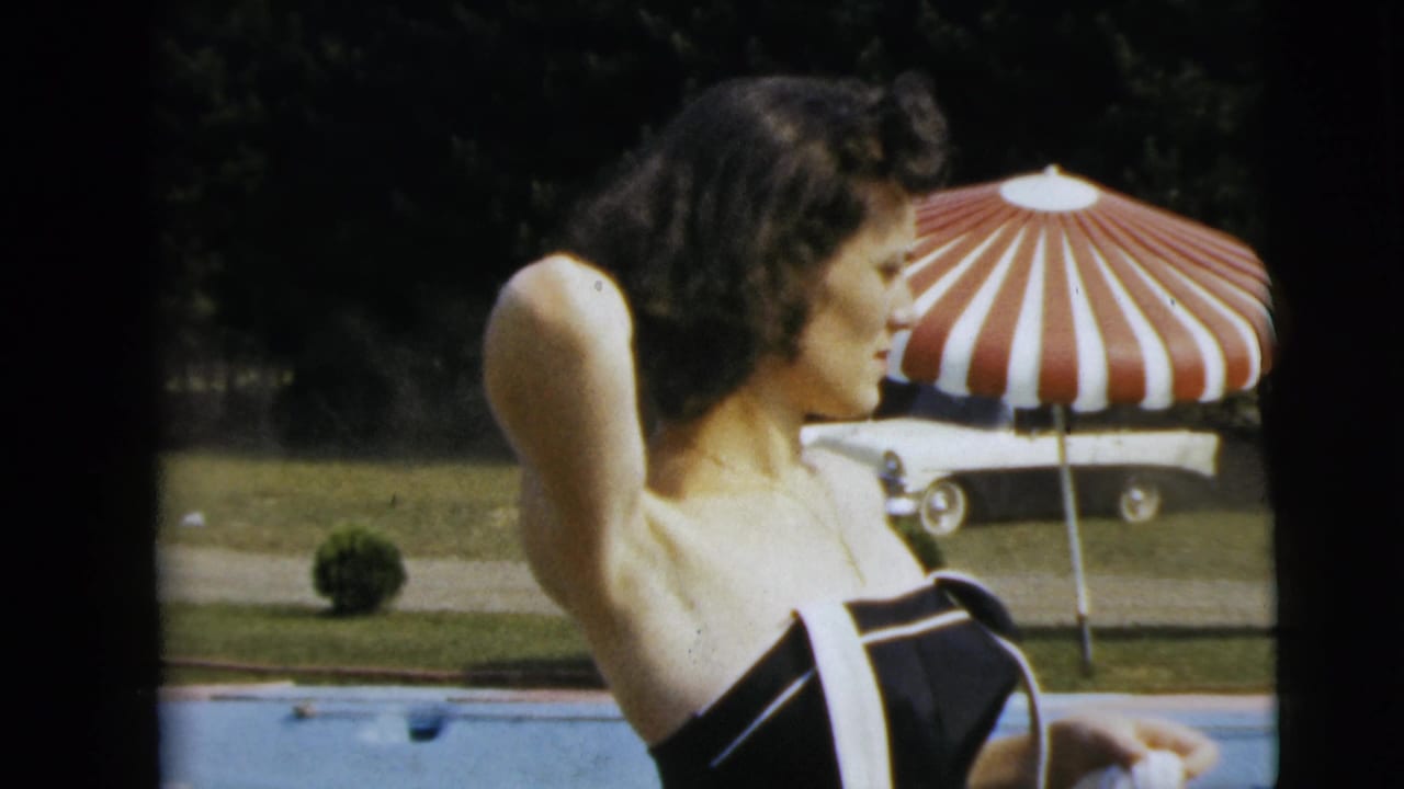 In Camden, New Jersey, 1962 home-movie footage, a woman in a black and white swimsuit stands by a pool near a striped umbrella.
