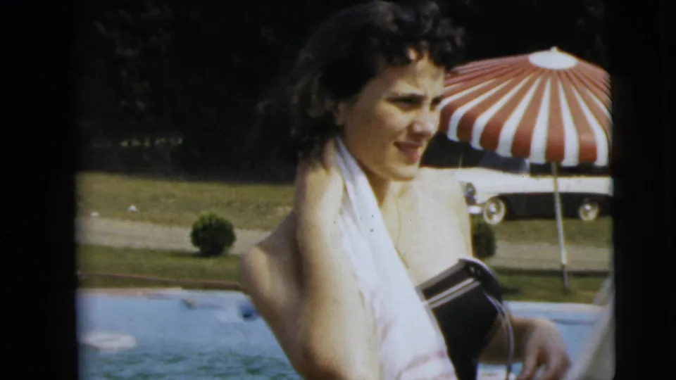 A woman in 1962 Camden poolside footage holds a white towel over her shoulder, with the pool, umbrella, lawn, and car behind her.