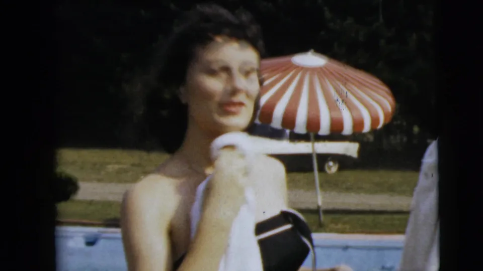 The woman dries herself with a white towel in soft 8mm footage, with a striped umbrella and poolside lawn visible behind her.