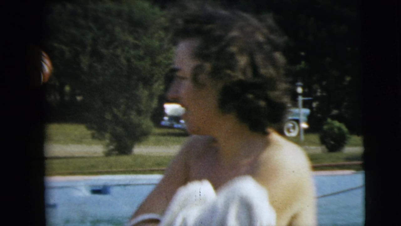 Woman in a white swimsuit smiling near an outdoor pool, with a parked older car behind her on the lawn.
