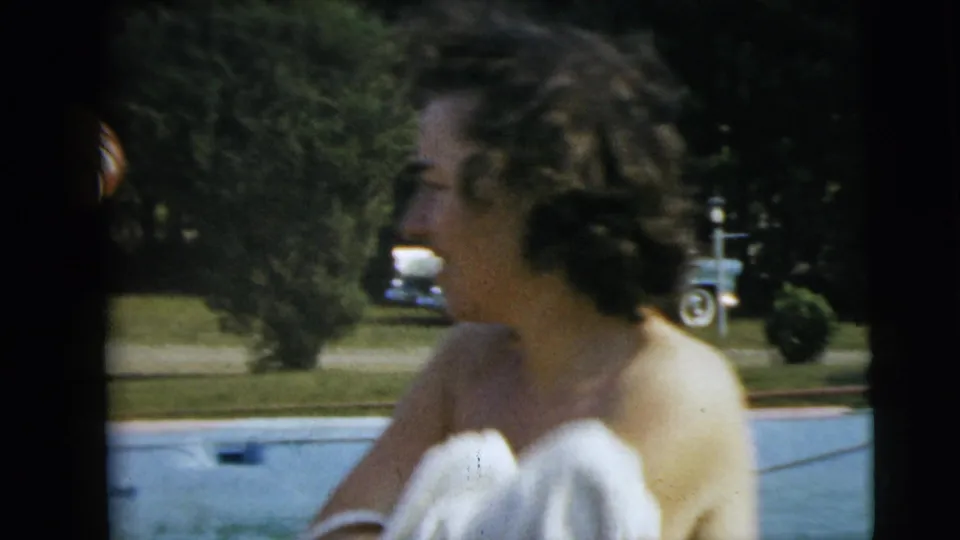 1962 Camden, New Jersey home-movie frame showing a smiling woman in a white swimsuit beside a pool, with lawn and an older car in the background.