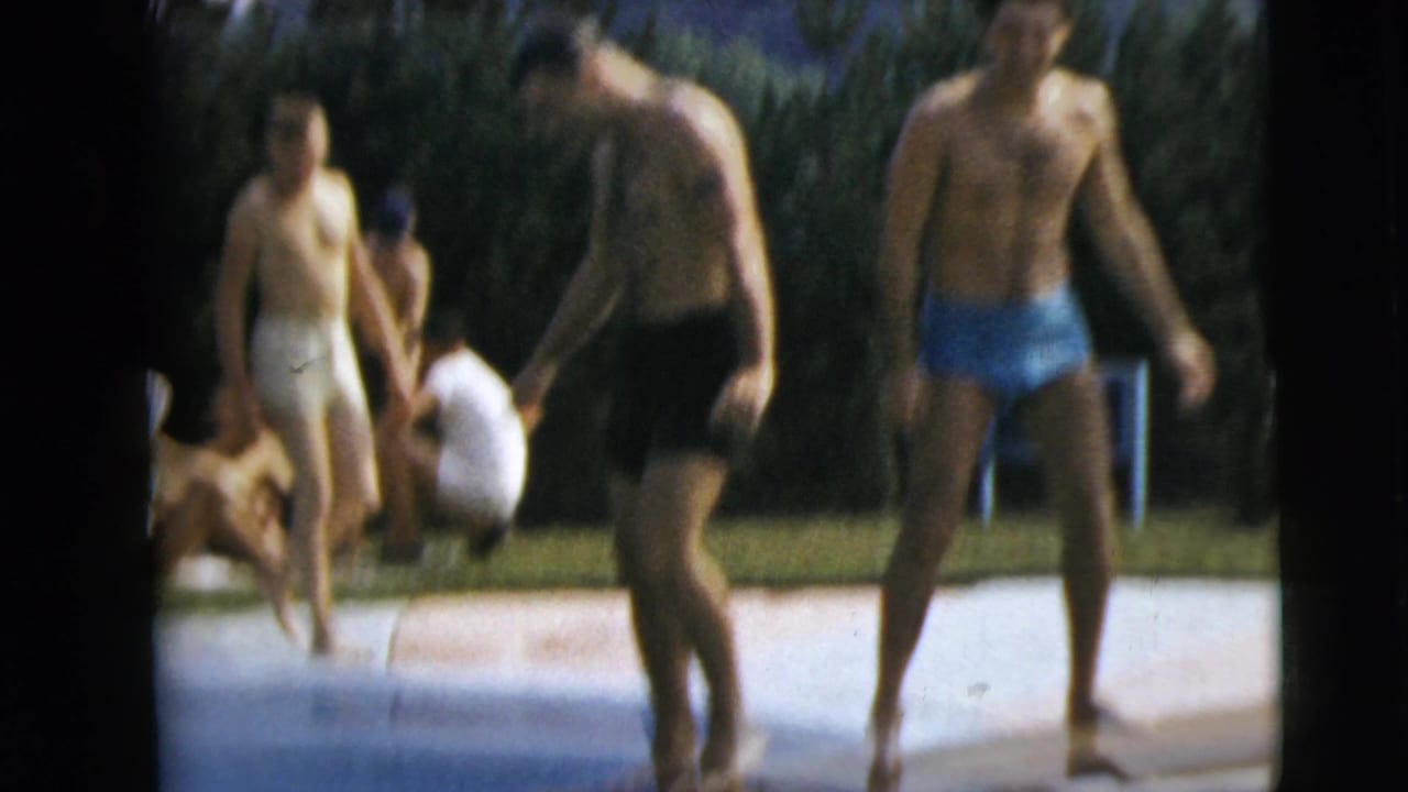 Men in swim trunks walk and stand near the edge of an outdoor swimming pool on a sunny lawn.