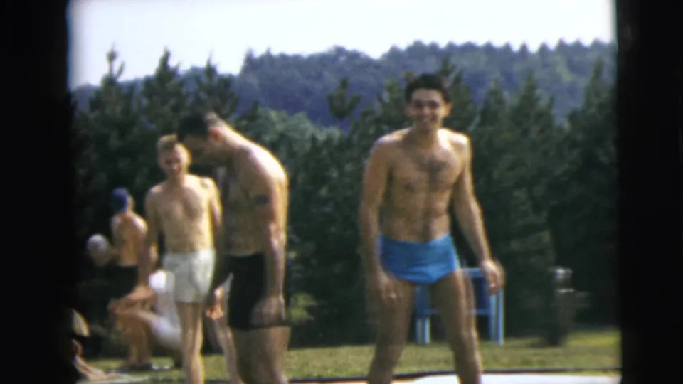 1962 Camden, New Jersey home-movie frame showing men in light, dark, and blue swim trunks beside a pool with seated adults behind them.