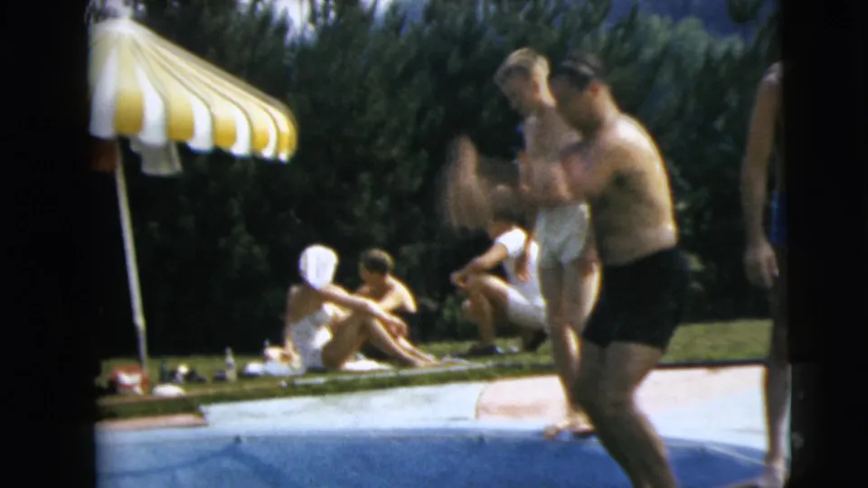 1962 Camden, New Jersey poolside frame with adults relaxing on grass, men near the water, and a yellow and white striped umbrella overhead.
