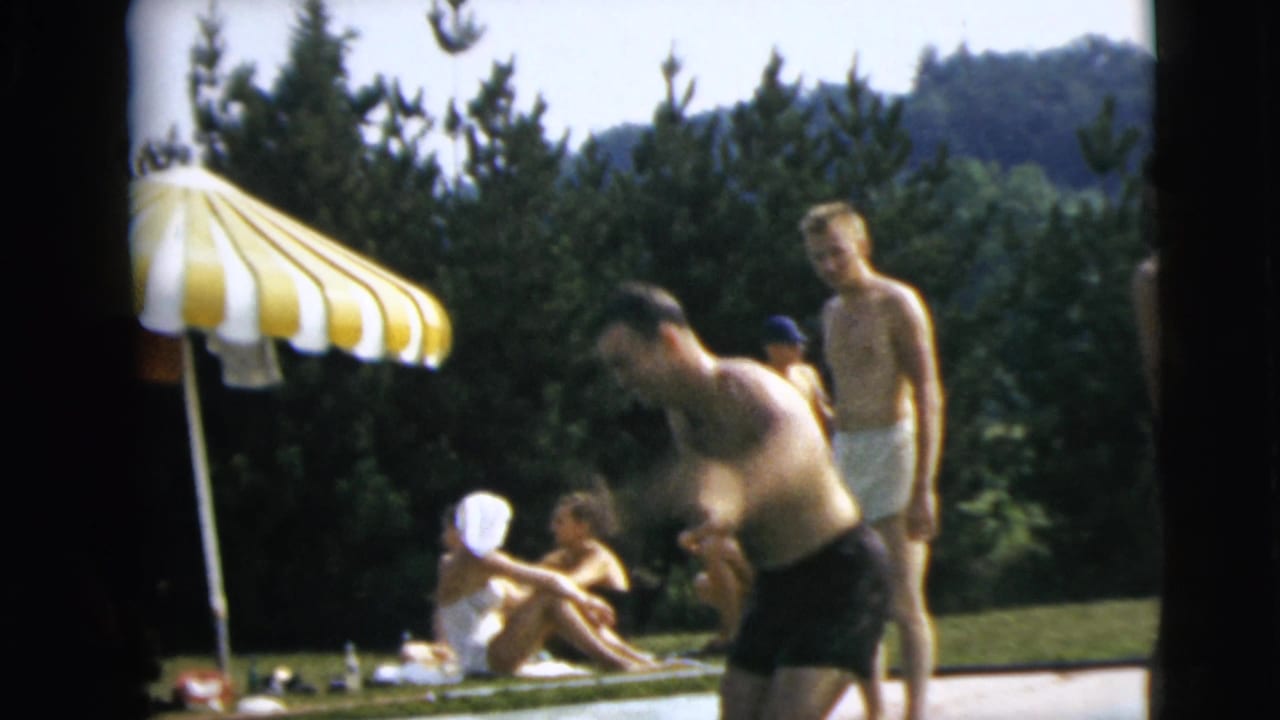People gather on grass beside a swimming pool under a striped umbrella.