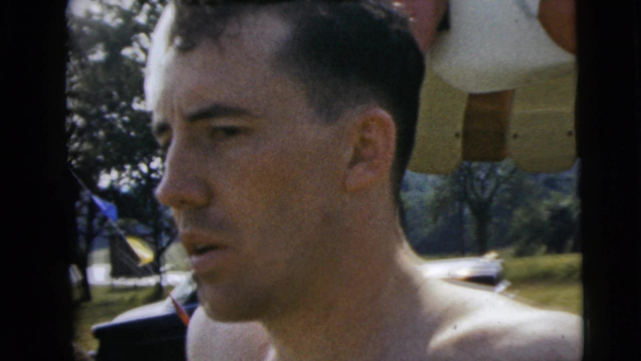 Close-up from 1962 Camden pool footage showing a shirtless man beside the pool area, with trees and umbrella fabric behind him.