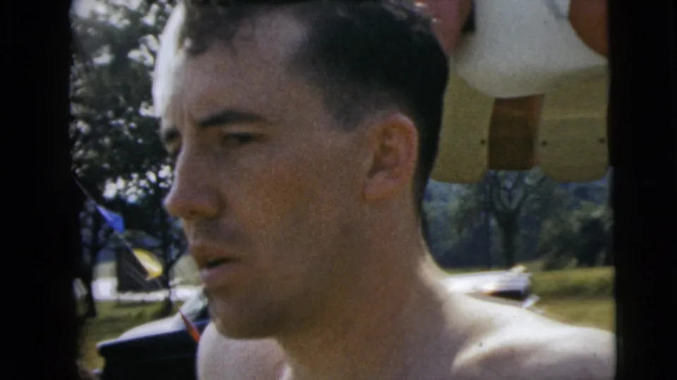 Close-up from 1962 Camden pool footage showing a shirtless man beside the pool area, with trees and umbrella fabric behind him.