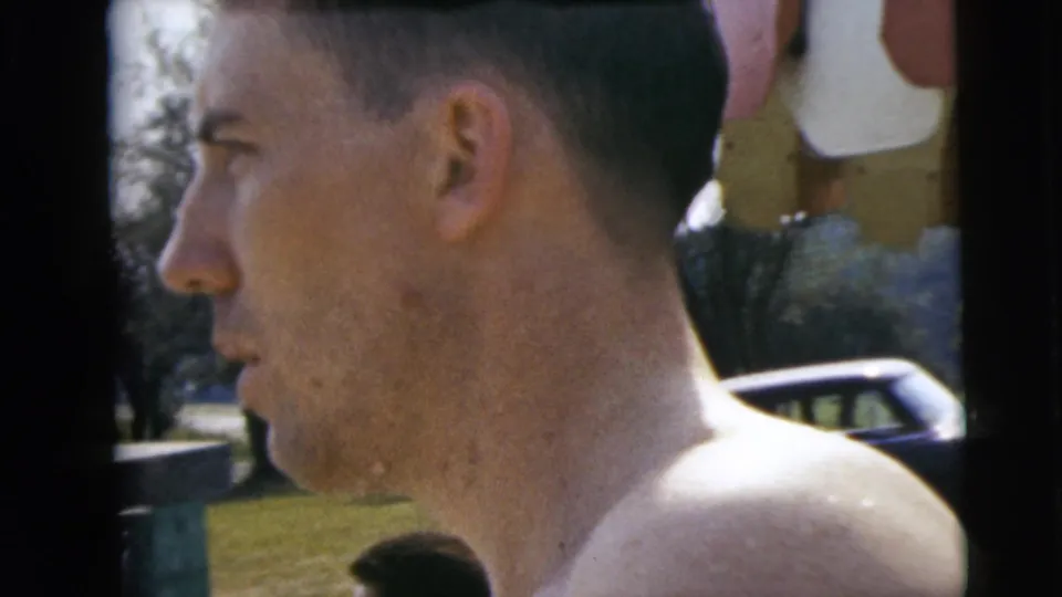 Profile close-up in the 1962 Camden home movie, showing a shirtless man with a parked car and poolside lawn behind him.