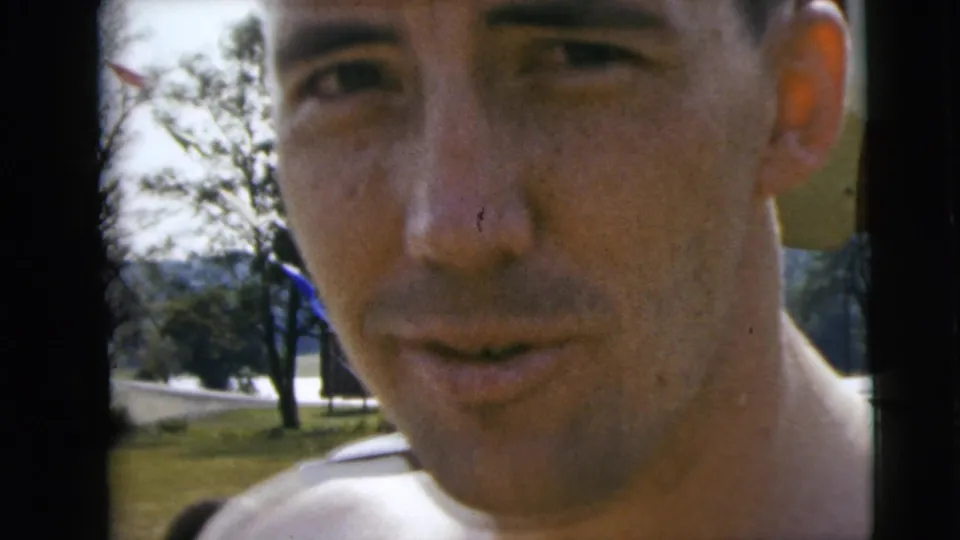 Close-up of a man in swimwear during 1962 Camden, New Jersey home-movie footage, with trees and water visible behind him.
