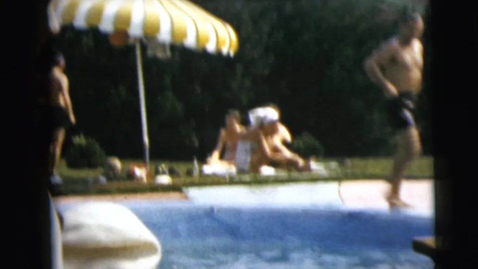 1962 Camden poolside home-movie frame with swimmers, a striped umbrella, lawn, trees, and blue pool water in bright daylight.