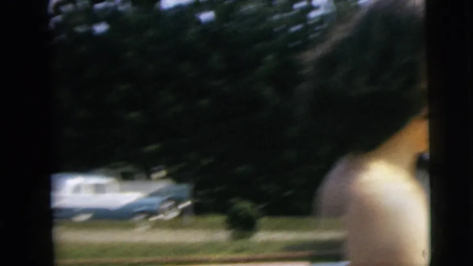 Blurred 1962 Camden home-movie frame with a shirtless person at frame edge, trees, and a parked blue-and-white car beyond the pool area.