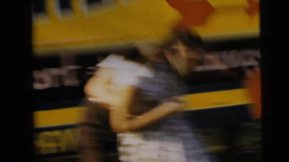 Blurred 1962 home-movie frame showing two people embracing near a yellow sign or vehicle with partially visible lettering.