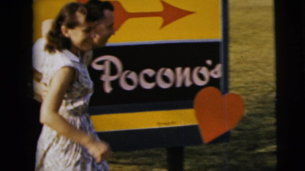 In Camden, New Jersey, 1962 home-movie footage, a woman and man stand near a yellow-and-black sign with partial lettering and a heart cutout.