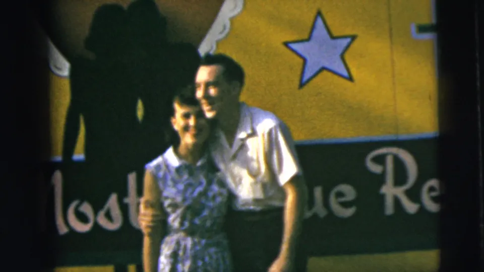 The couple remains arm in arm before a yellow-and-black sign, with a large shadow crossing the background in this 1962 Camden home-movie frame.