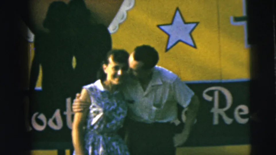In the 1962 Camden home movie, the man leans in toward the woman's cheek as she smiles near the yellow and green sign.