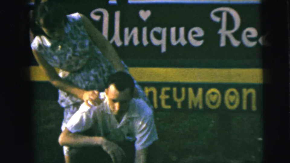 The woman in a blue patterned dress begins climbing onto the man's back during the playful 1962 Camden home-movie moment.