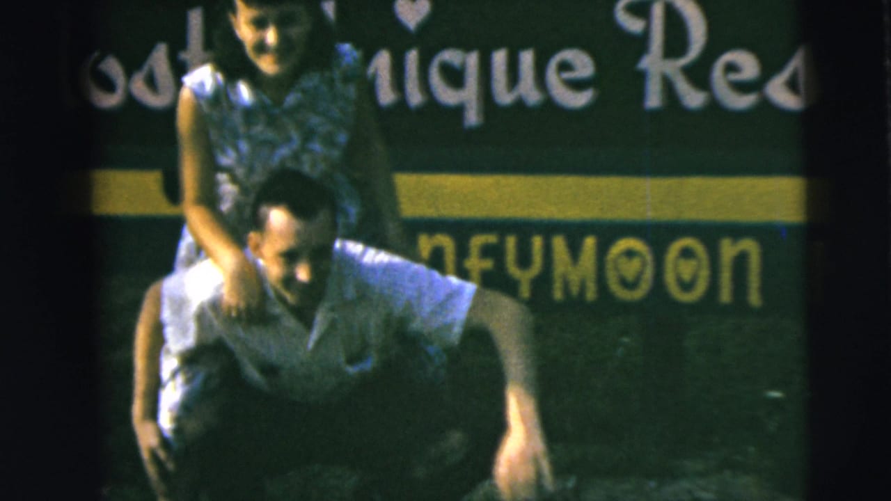 1962 home-movie frame from Camden, New Jersey, showing a smiling couple in a piggyback pose before a green honeymoon resort sign.