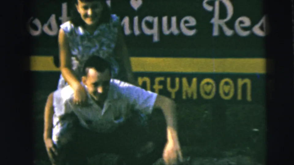1962 home-movie frame from Camden, New Jersey, showing a smiling couple in a piggyback pose before a green honeymoon resort sign.