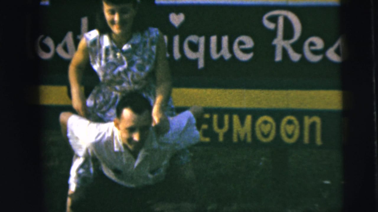 Home-movie still of a couple smiling during a piggyback ride, with honeymoon sign lettering visible behind them.