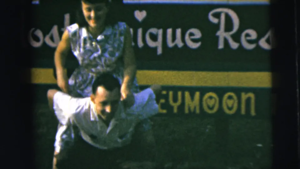 Home-movie still of a couple smiling during a piggyback ride, with honeymoon sign lettering visible behind them.