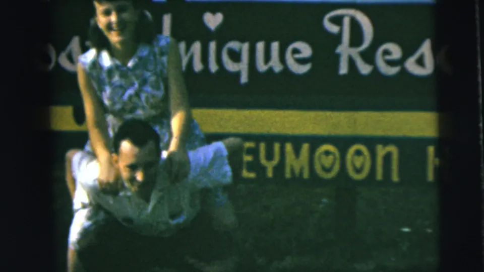 The woman smiles toward the camera while the man carries her piggyback before a green sign with partial honeymoon text.