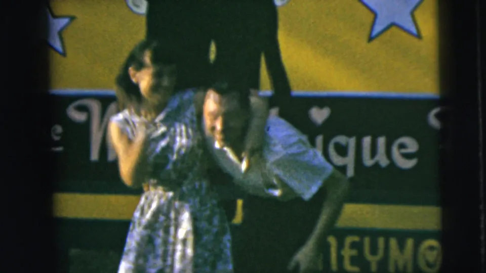 1962 Camden home-movie frame of the couple mid-dance, partly blurred, with a star-decorated green and yellow backdrop behind them.