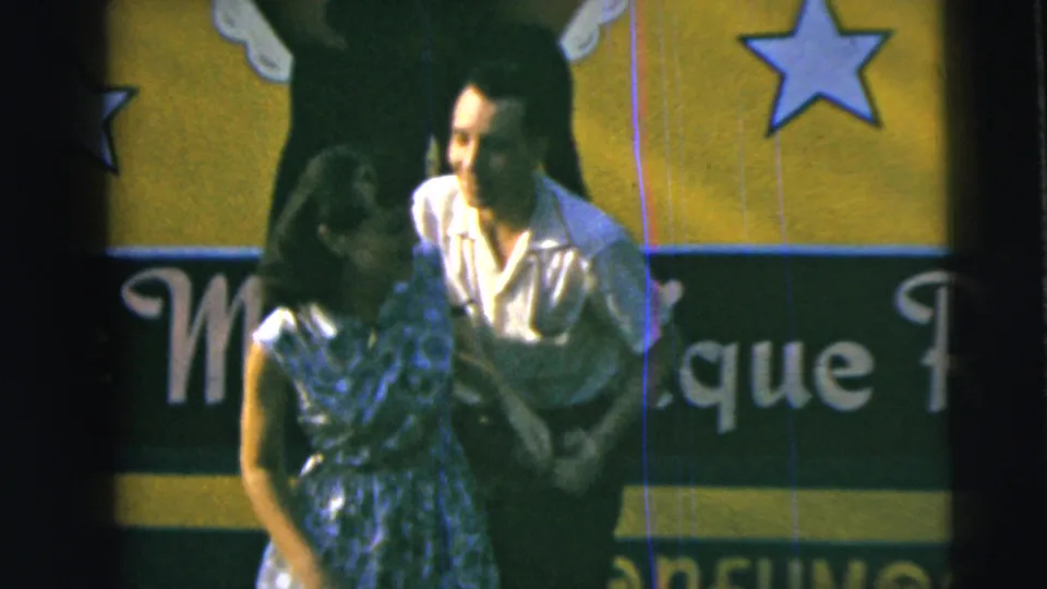 1962 Camden frame showing a man smiling at his partner as they hold hands near partial lettering and star shapes on the backdrop.