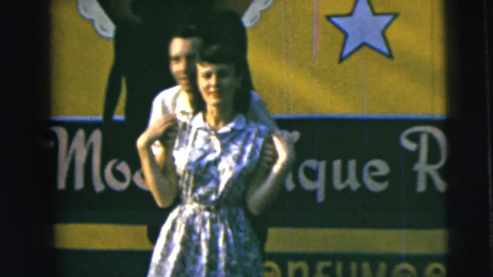 In 1962 Camden home-movie footage, a young couple stands close together before a yellow and green sign with a star and partial lettering.