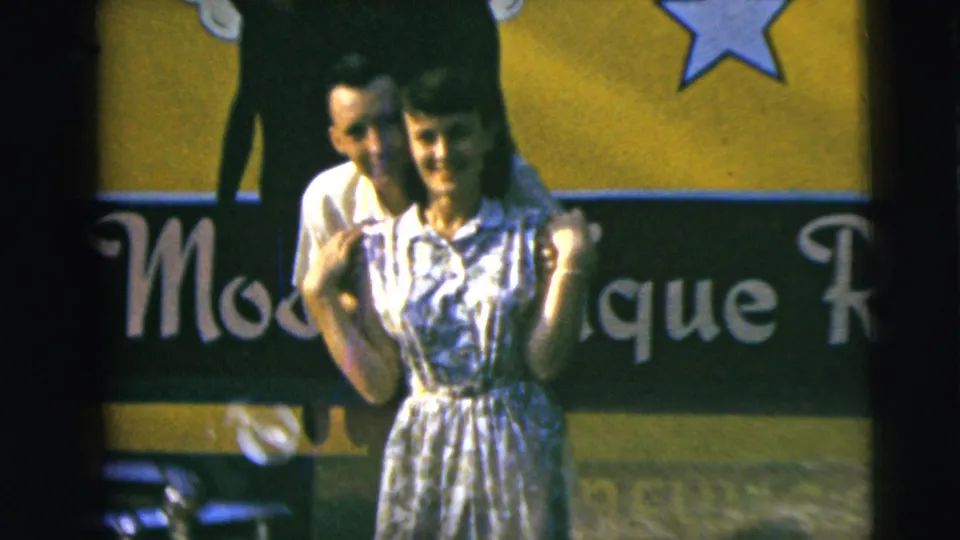 A woman in a blue and white patterned dress stands before the sign while a man leans close behind her in this Camden home-movie frame.