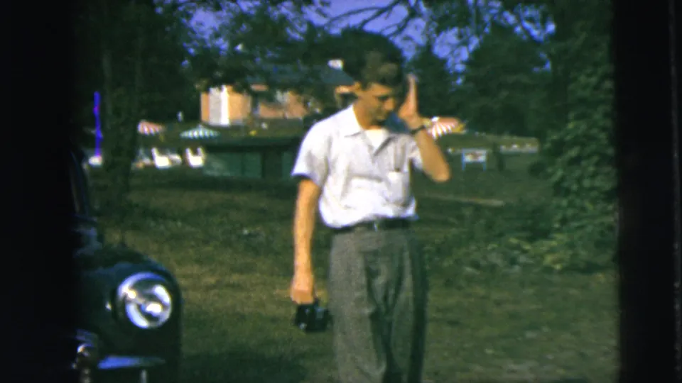 A man in a light shirt walks outdoors holding a camera, with grass, trees, buildings, and part of a parked car visible behind him.