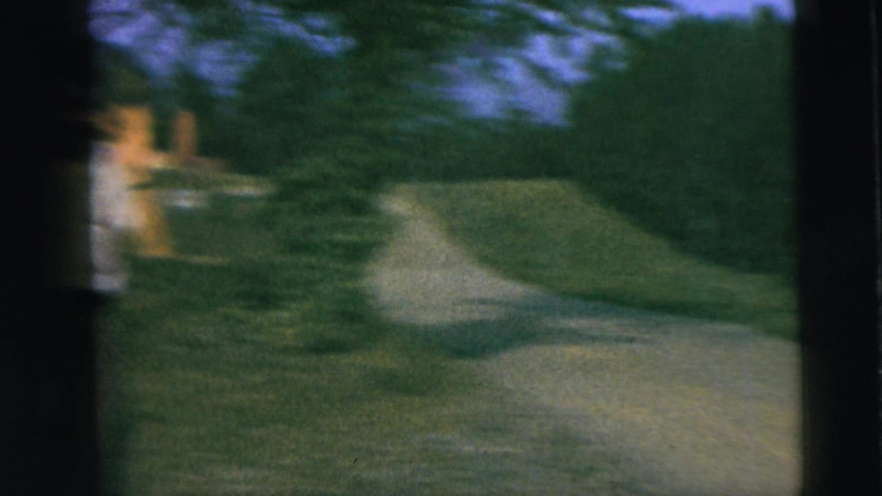 1962 Camden home-movie frame with a soft, blurred view of grass, trees, a path, and a woman near the left edge.
