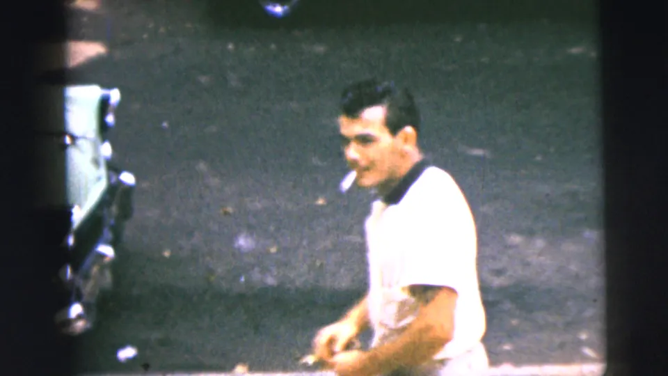 Brief 1962 Camden home-movie glimpse of an adult man in a white short-sleeve shirt holding a cigarette near a street surface and partial vehicle edge.