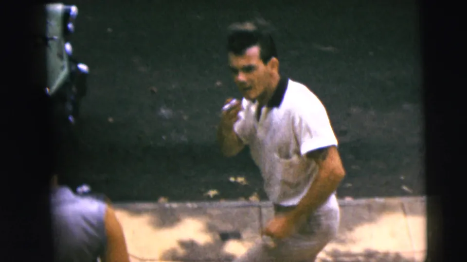 In 1962 Camden home-movie footage, a man in a white polo shirt smokes while standing near a street edge.