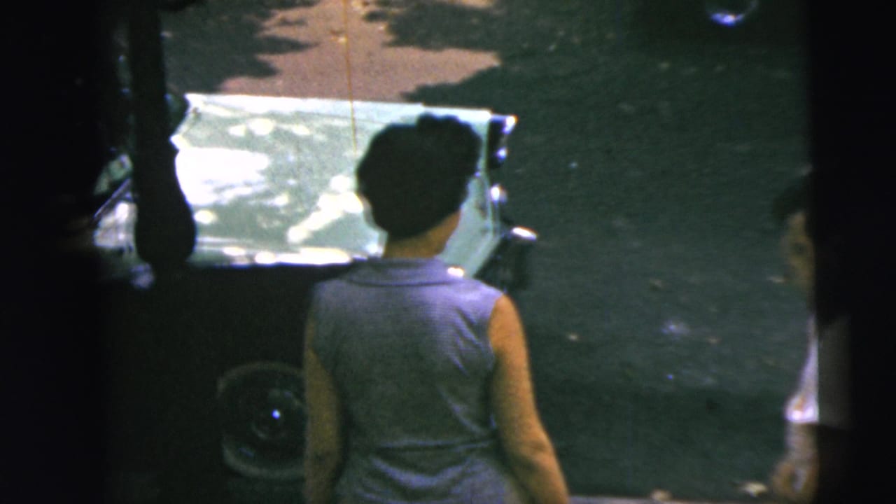 A woman stands with her back partly turned near a green car with chrome trim in the 1962 Camden street scene.
