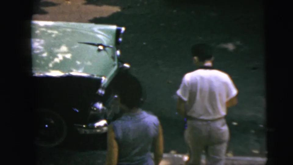The man and woman are seen from behind near a parked green car in candid 1962 Camden home-movie footage.
