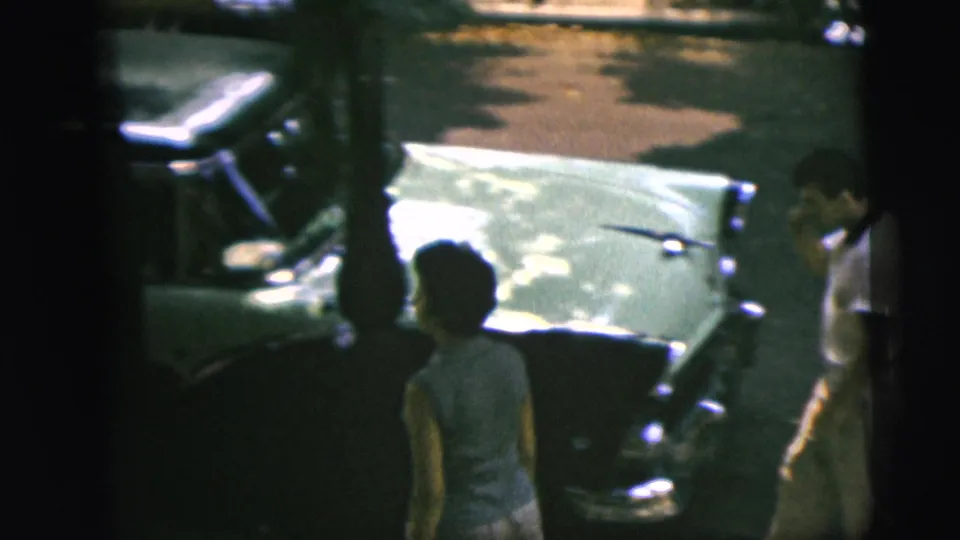 A woman stands by the green car as a man in a white shirt appears at the frame edge in the 1962 Camden street footage.
