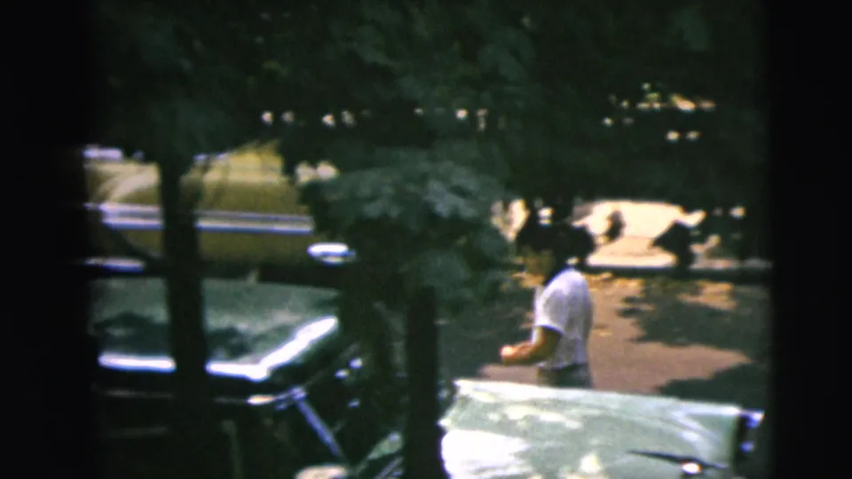 A man in a light shirt stands beside the two-tone sedan on a shaded Camden street, with parked cars and trees in the background.
