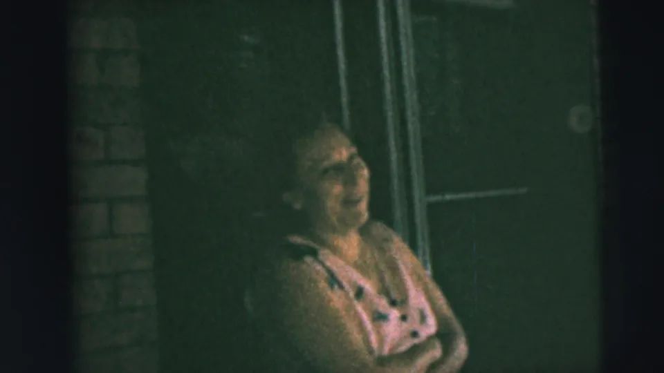 In Camden, New Jersey home-movie footage from 1962, a woman in a pink dress laughs on a dim porch near a brick wall and door.