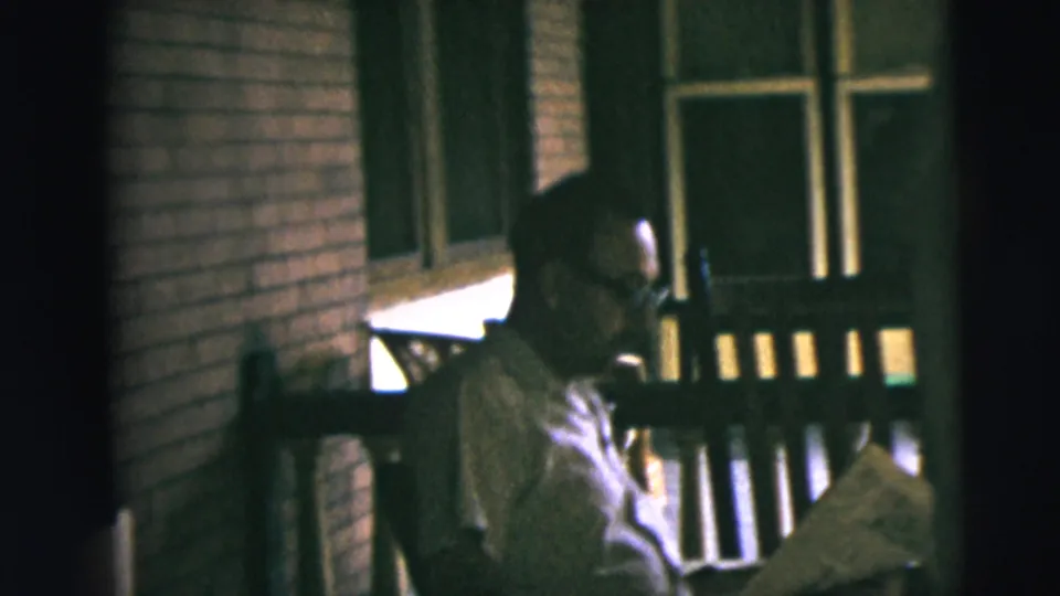 A man with glasses sits on a wooden porch bench in Camden, New Jersey, reading a newspaper in dim 1962 home-movie footage.