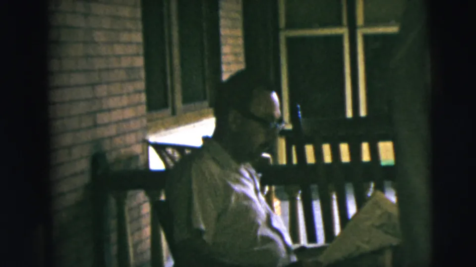 The man remains seated on the porch bench with a newspaper, framed by brick walls, a window, and porch railings in the 1962 Camden footage.