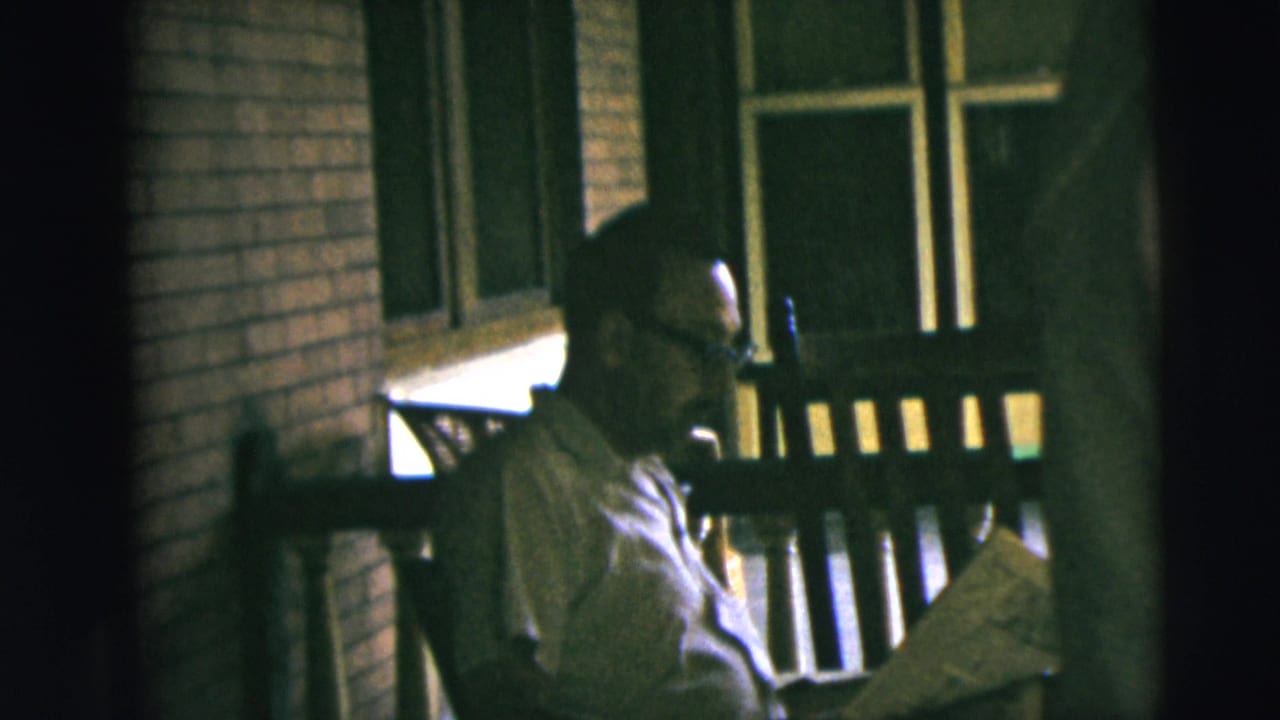 1962 Camden home-movie frame showing a man in glasses seated in a wooden rocking chair on a porch, looking down at a newspaper.
