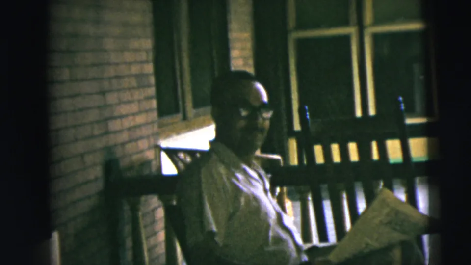 1962 Camden home-movie frame showing the man facing the camera from a wooden rocking chair with a newspaper across his lap.