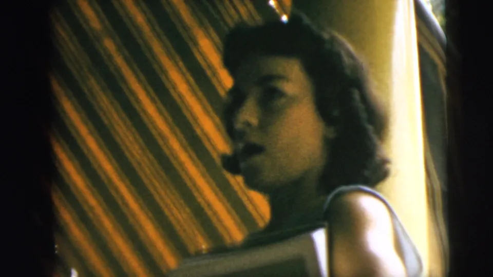 Young woman in profile near a striped awning in 1962 Camden home-movie footage, with soft 8mm exposure and shadow.