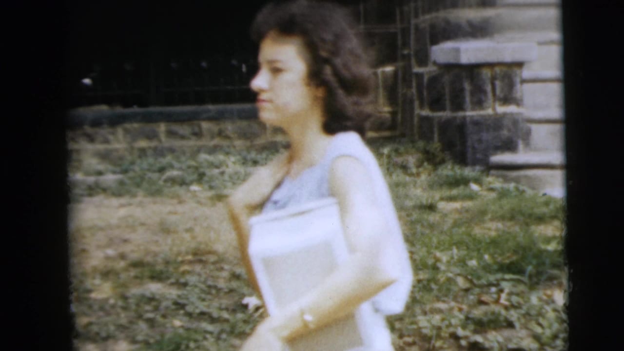 Young woman in a light sleeveless dress walking outdoors near a stone building and grass.