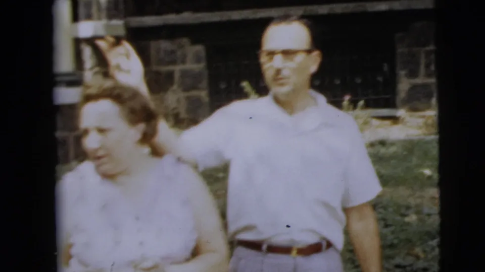 Man in glasses and a white shirt appears to gesture or speak behind a woman outdoors in the supplied 1962 Camden footage.