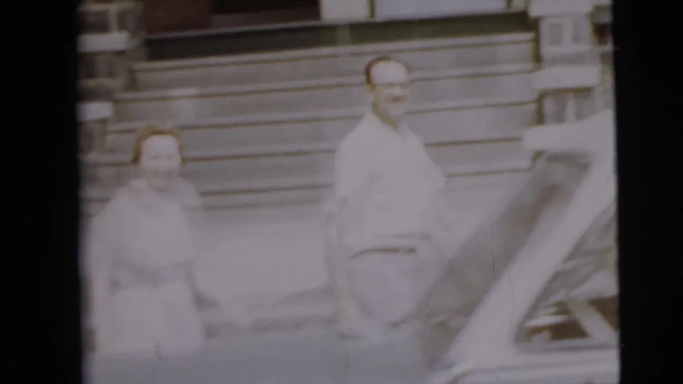 In 1962 Camden home-movie footage, a man and woman stand near house steps with a white car in the foreground.