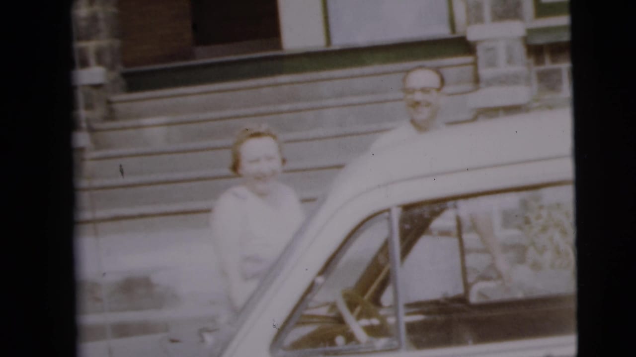 Woman and man smile beside a parked car outside a house.