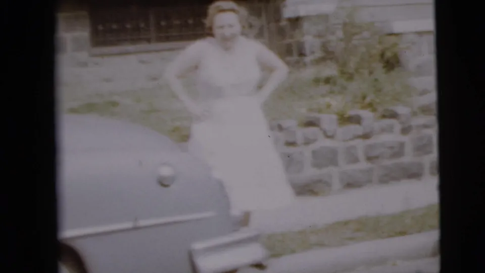 A woman in a light dress stands near the front of a white car outside a Camden residence in faded 8mm footage.