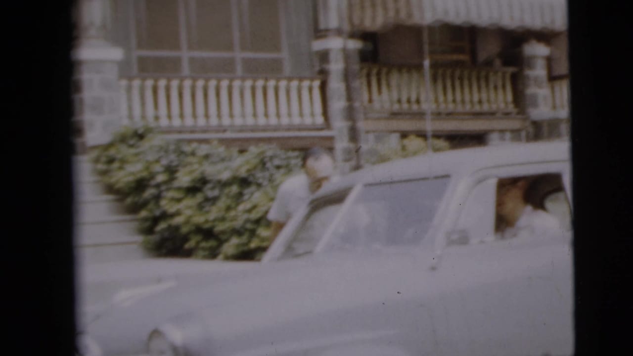 Man standing near an older sedan outside a residential building.